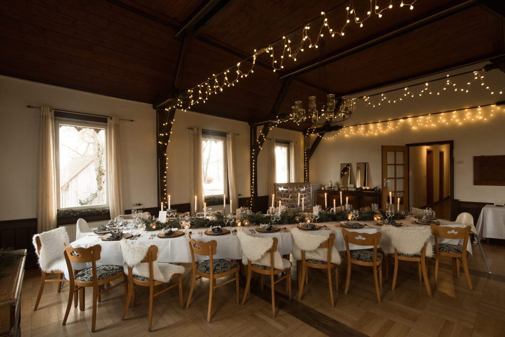 Eingedeckte Tafel für winterliche Hochzeitsfeier mit Lichterketten über der Tafel und Kerzen, Holzplatten und Teelichtern auf dem Tisch.