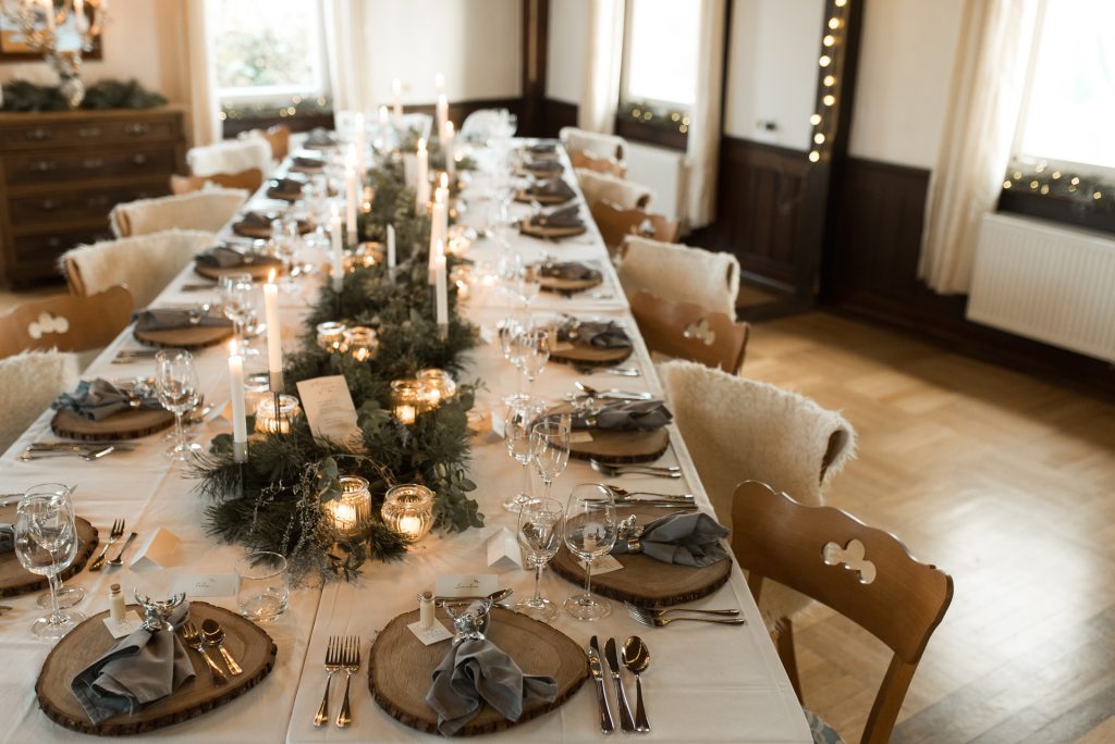 Winterlich rustikal eingedeckte Tafel in einem hellen Raum mit Lichterketten und Kerzenbeleuchtung.